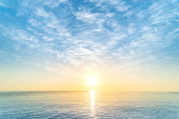 Bright sun rising over the ocean under a soft blue sky